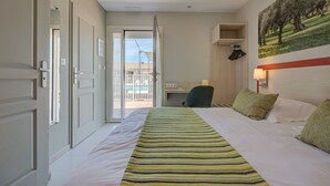 A hotel room with a bed, a desk, and a chair. There is a door leading to a balcony with a view of buildings and a pool. - Hotel Saint Louis (Le Thor)