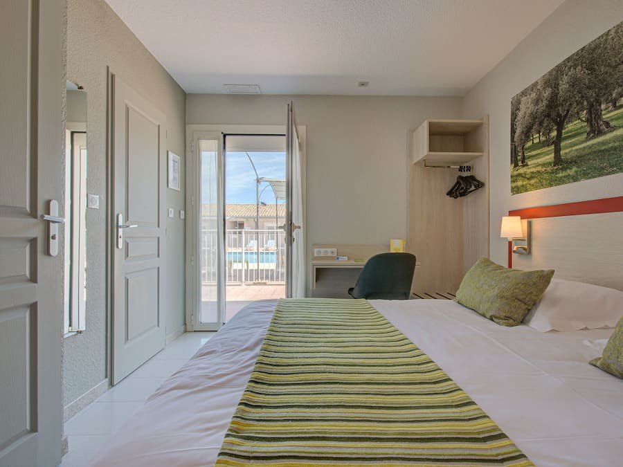 A hotel room with a bed, a desk, and a chair. There is a door leading to a balcony with a view of buildings and a pool.