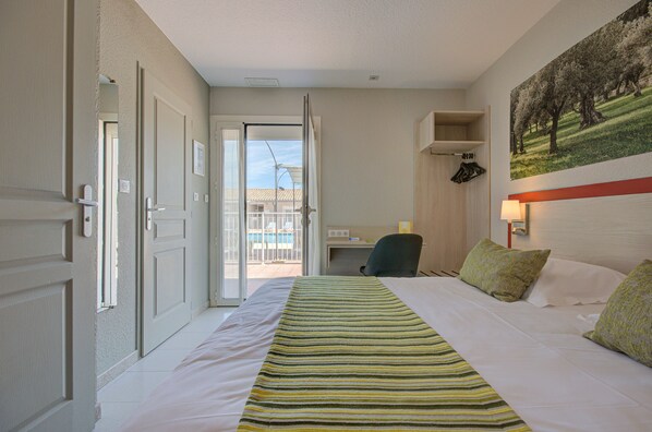 Habitación de hotel con cama, escritorio y silla. Se ve una puerta que da a un balcón con vistas a edificios y una piscina.