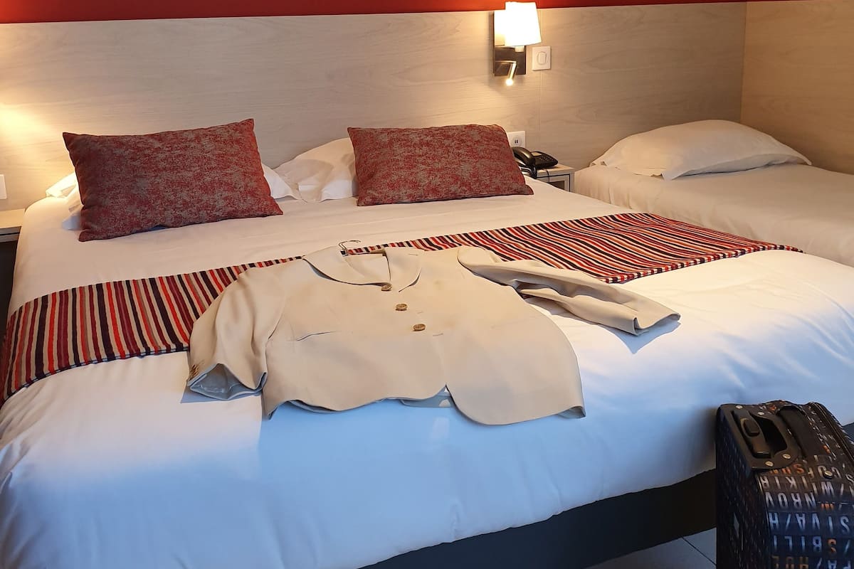 A hotel room with a bed, two pillows, a bedside table with a lamp, a telephone, and a suitcase.
