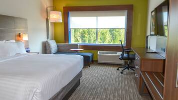 A hotel room with a large bed, a desk, a chair, a sofa, a window with a view of trees, and a flat-screen TV.