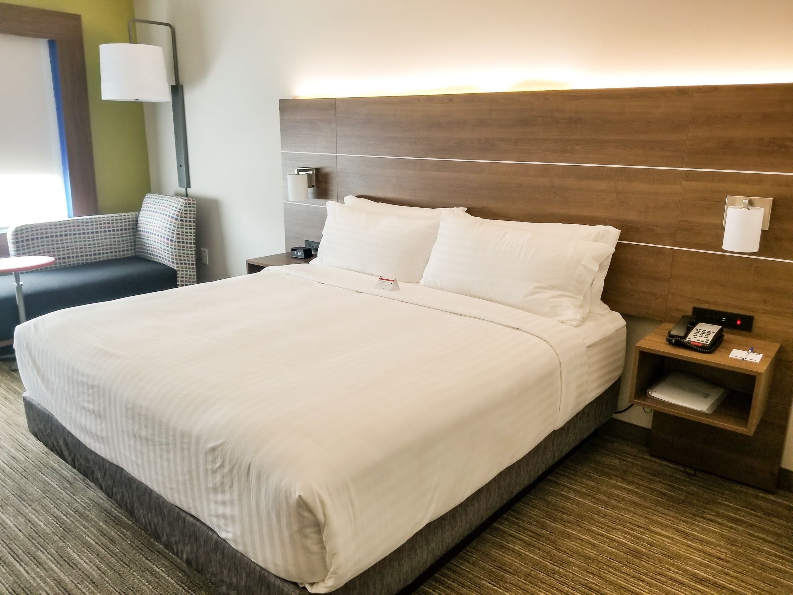 Hotelkamer met een groot bed, een nachtkastje met telefoon en lamp, een klein tafeltje met stoel en een wandlamp.