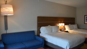 Quarto de hotel com sofá azul, duas camas com roupa de cama branca, cabeceira de madeira, mesa de cabeceira com abajur e tomada na parede.
