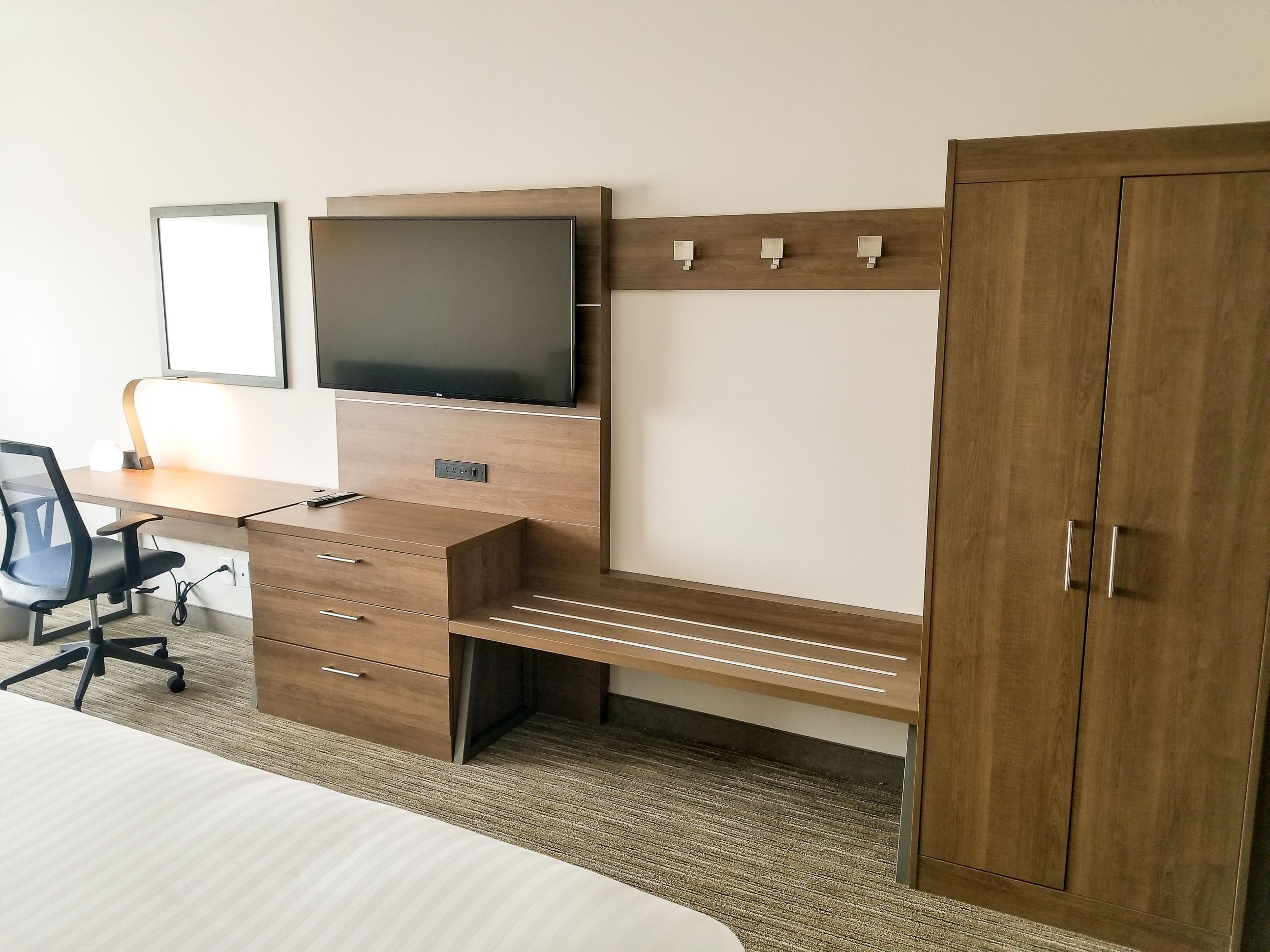 A modern hotel room with a wooden desk, a flat-screen TV, a wardrobe, and a bed.