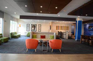 A modern lobby with a sofa, two orange chairs, a coffee table, and a reception desk. - Holiday Inn Express & Suites Charlotte NE - University Area by IHG (Charlotte)