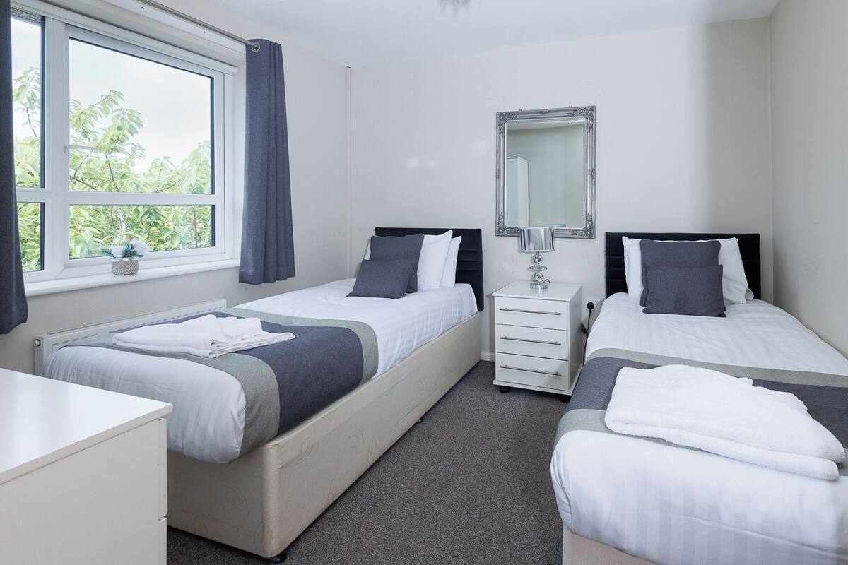 A twin room with two beds, a bedside table, a dresser, a mirror, and a window with curtains.