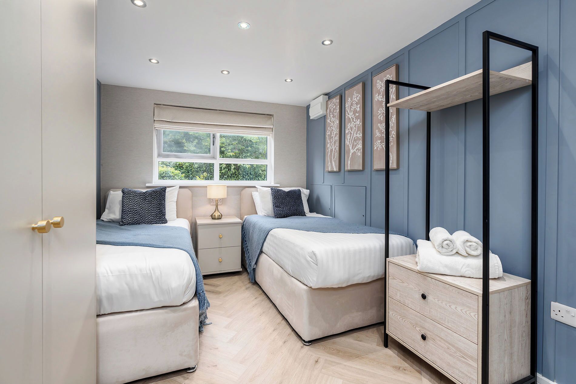 A bedroom with two beds, a window with a view of greenery, a bedside table with a lamp, and a mirror with a towel rack.