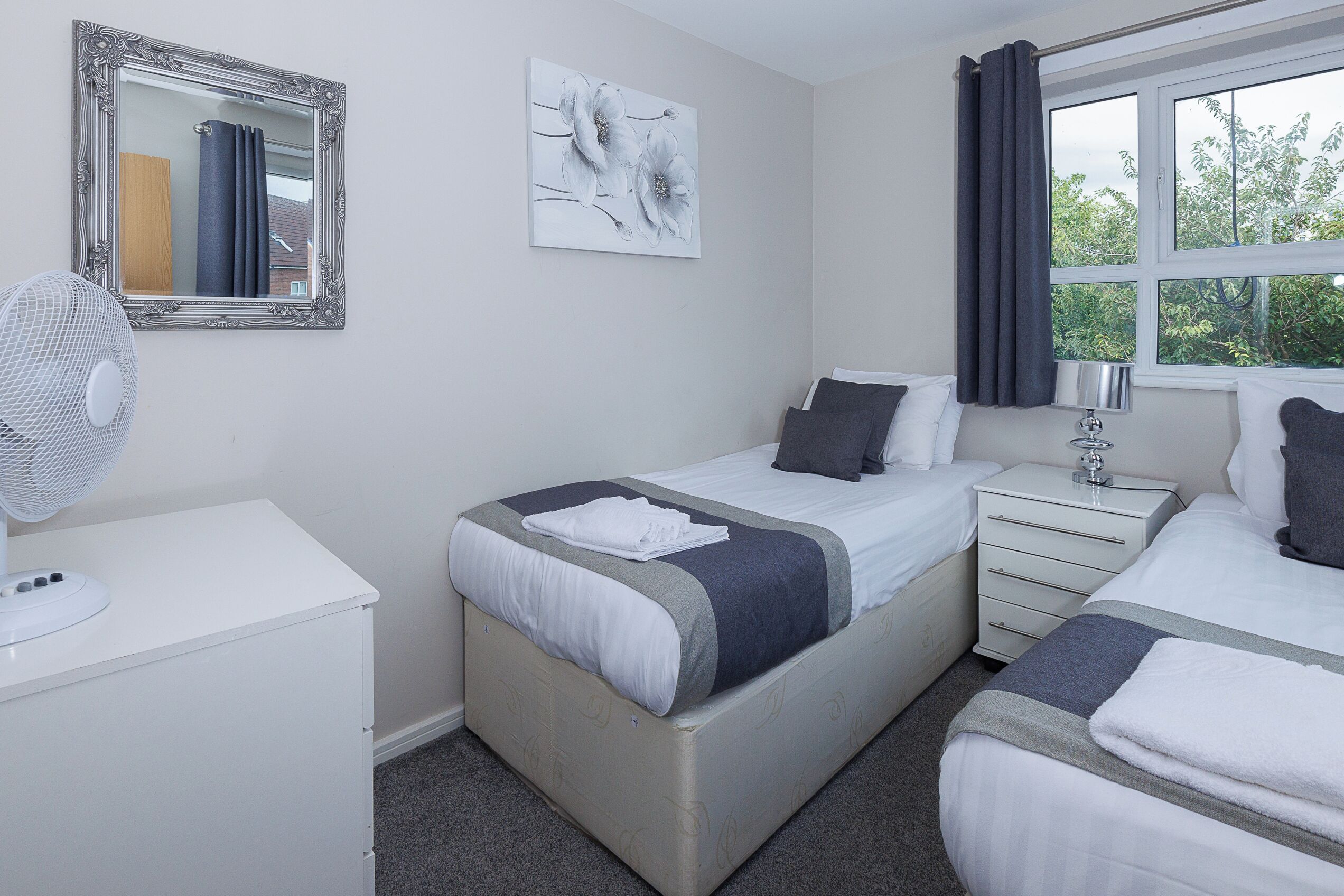 A hotel room with two beds, a bedside table, a nightstand, a mirror, and a window with curtains.