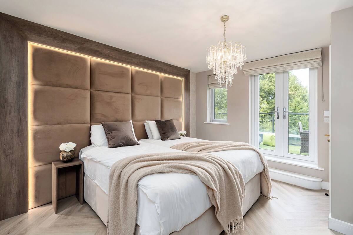 A modern bedroom with a large bed, a wooden headboard with illuminated panels, a chandelier, and a view of greenery through the windows.