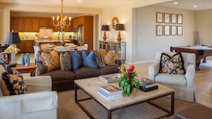 Amplia sala de estar con un sofá de cuero marrón, un sillón blanco y una mesa de centro con libros y una planta. Se ve una cocina con armarios de madera y un comedor con candelabro al fondo.