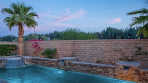 Una piscina con cascada, rodeada por un muro de ladrillo y palmeras.