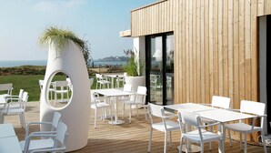 Een terras met witte stoelen en tafels, een houten gebouw met grote ramen, en uitzicht op de zee.