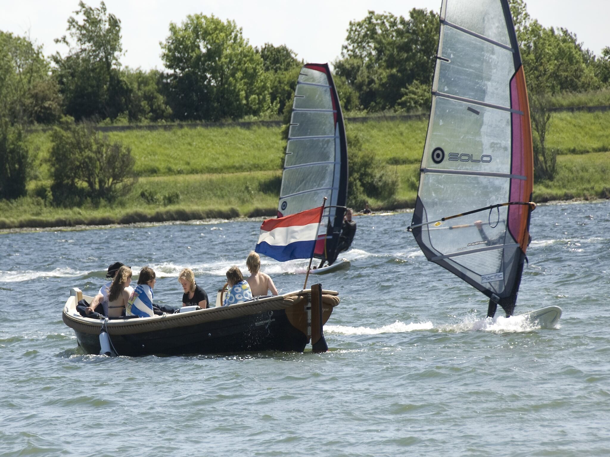 A boat with people on board, a sailboat with a flag, and a calm water body with greenery in the background.