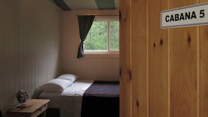 A single bed with white bedding in a room with wooden walls and a window with curtains. A sign on the wall reads "CABANA 5". - Godspeed Events & Lodging (Port Matilda)