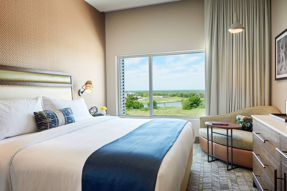 Ett hotellrum med en stor säng, en soffa, en byrå och utsikt över en sjö och grönområden.