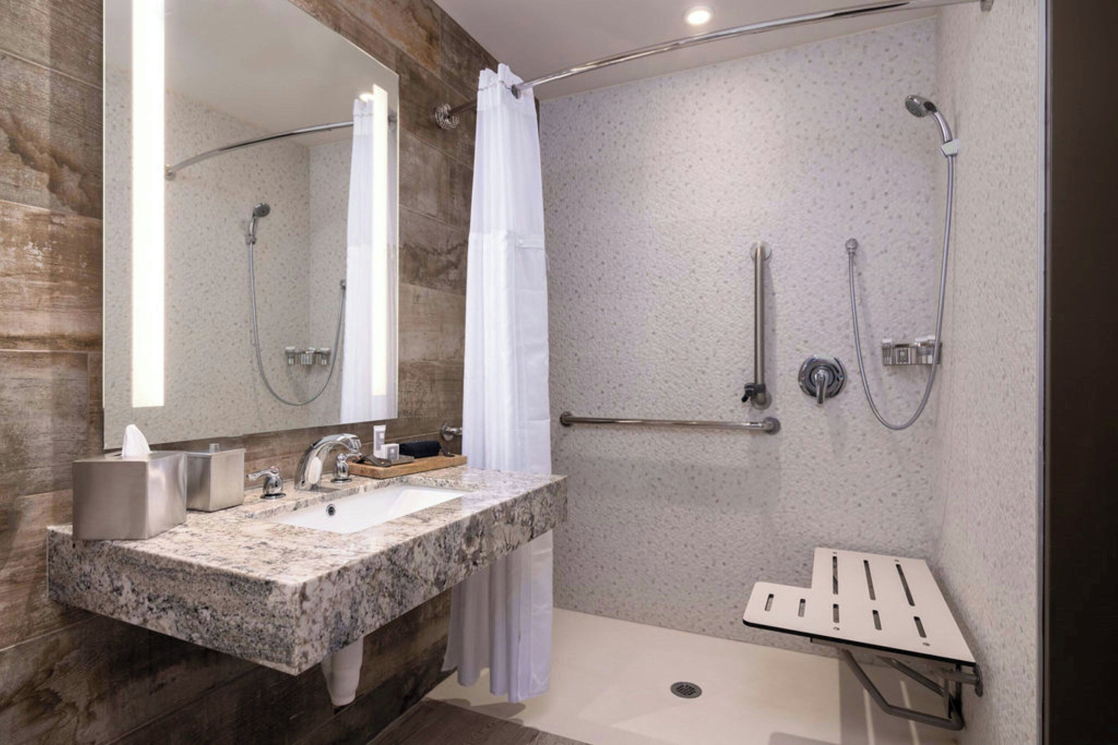 Un baño moderno con encimera de granito, un espejo grande, ducha con puerta de cristal y barra de apoyo.