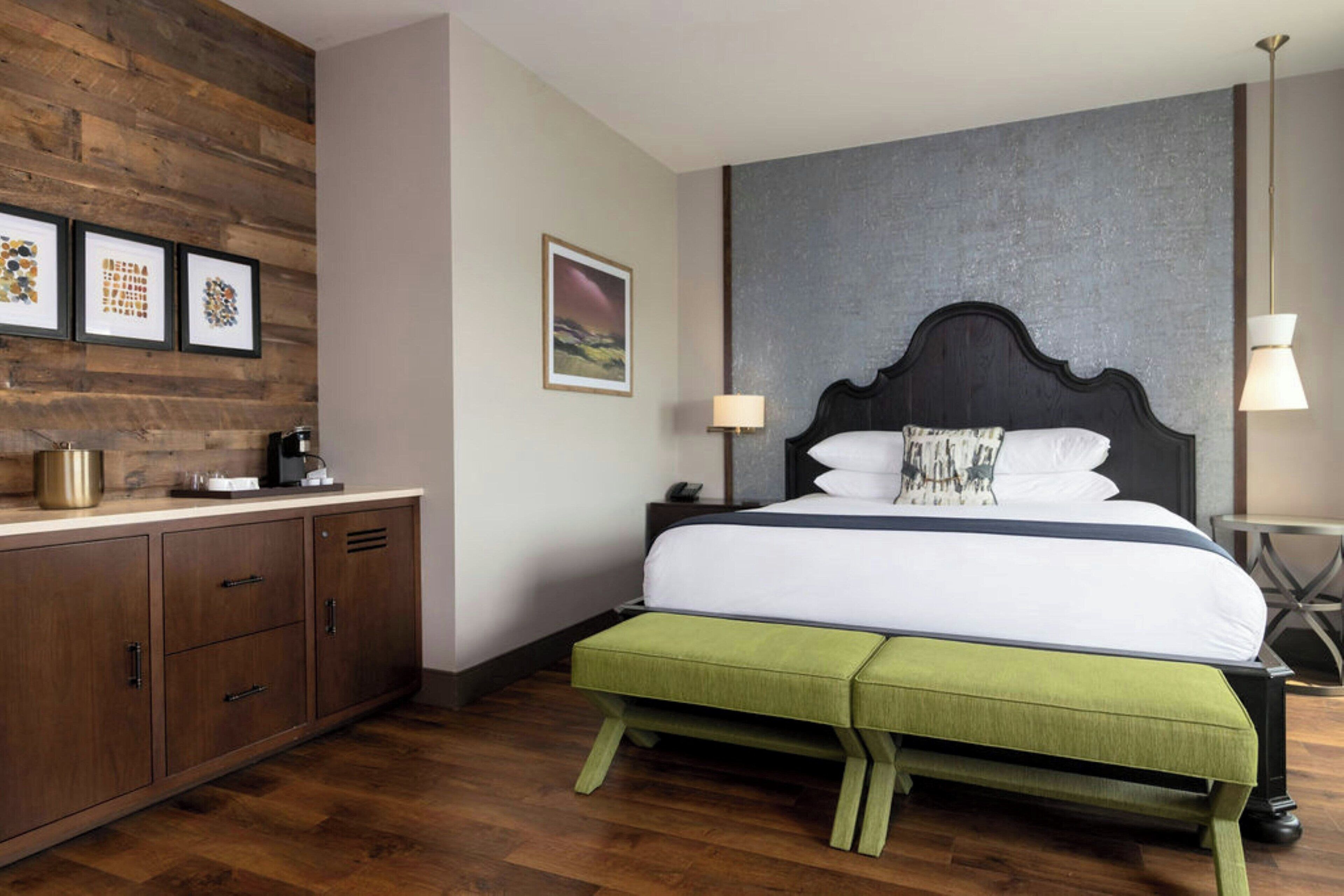 A bedroom with a wooden headboard, a green bench, a bed with white bedding, a nightstand, a lamp, and framed pictures on the wall.