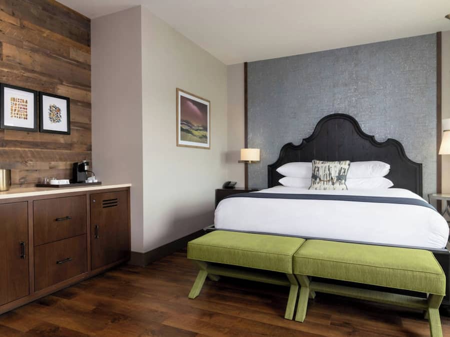 A bedroom with a wooden headboard, a green bench, a bed with white bedding, a nightstand, a lamp, and framed pictures on the wall.