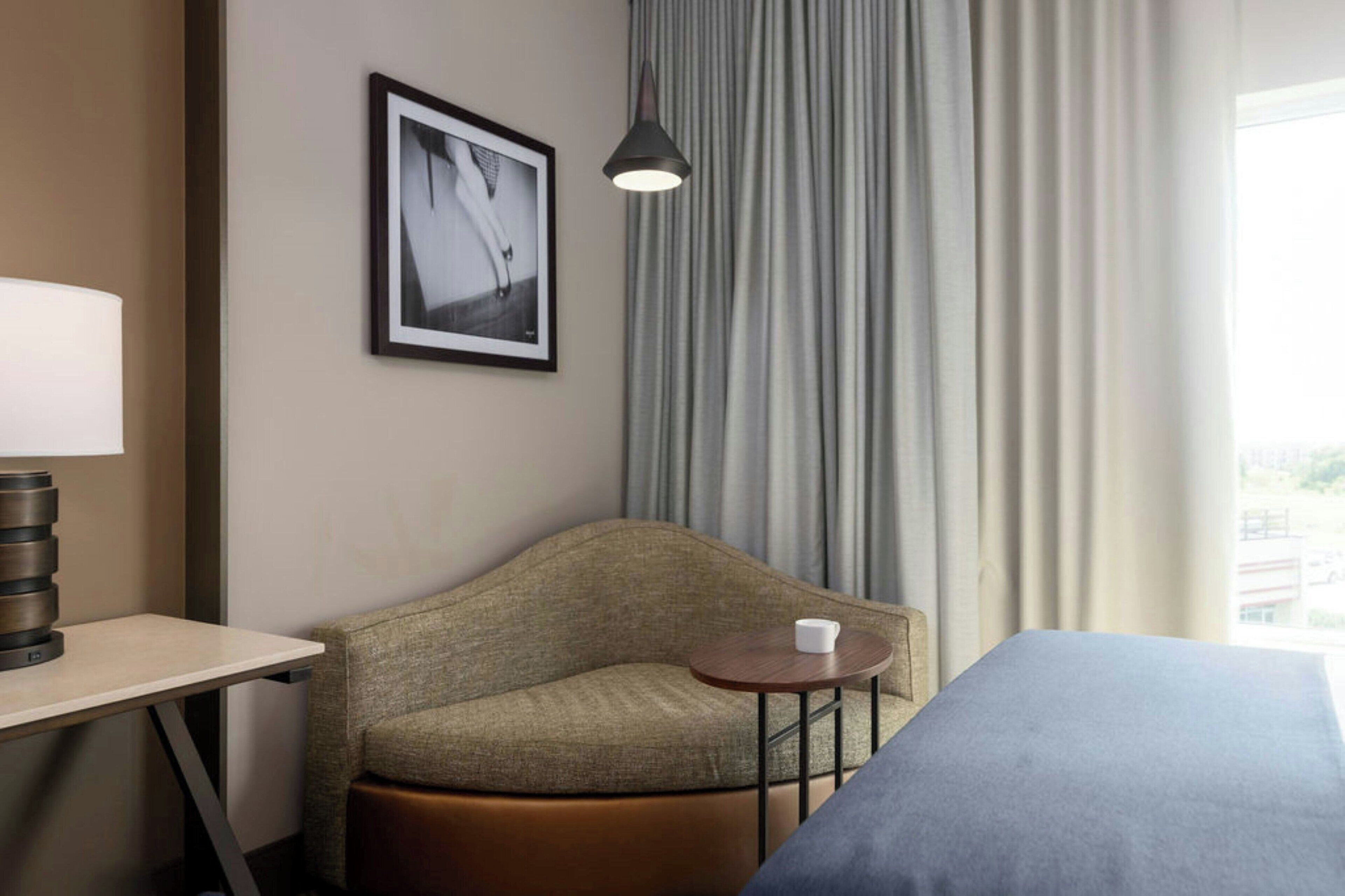 Et hotelværelse med en seng, en sofa, et lille bord med en kop, en lampe og et billede i ramme på væggen.