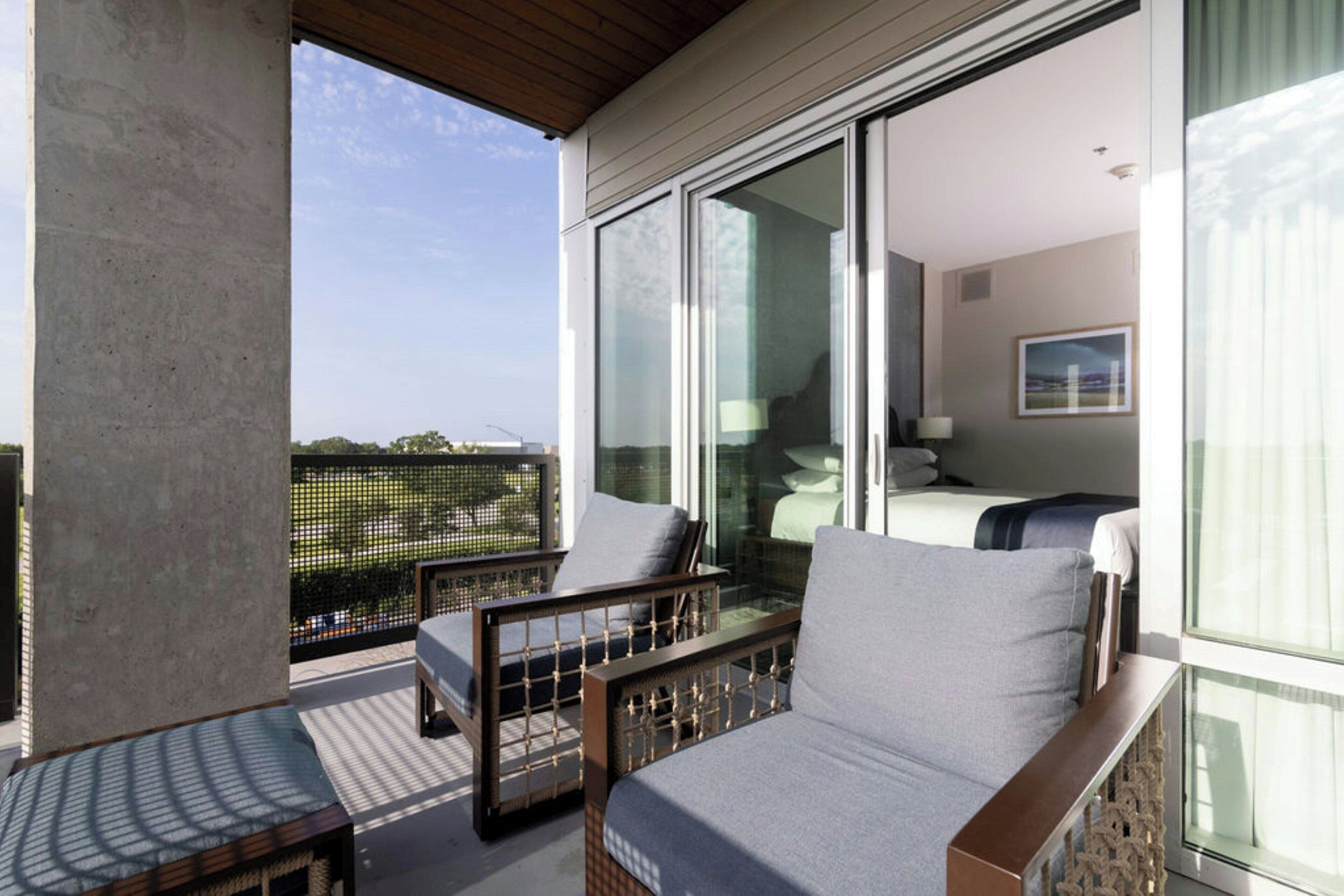 Un balcón con sofá y sillón, cama con vistas, y un ventanal grande.