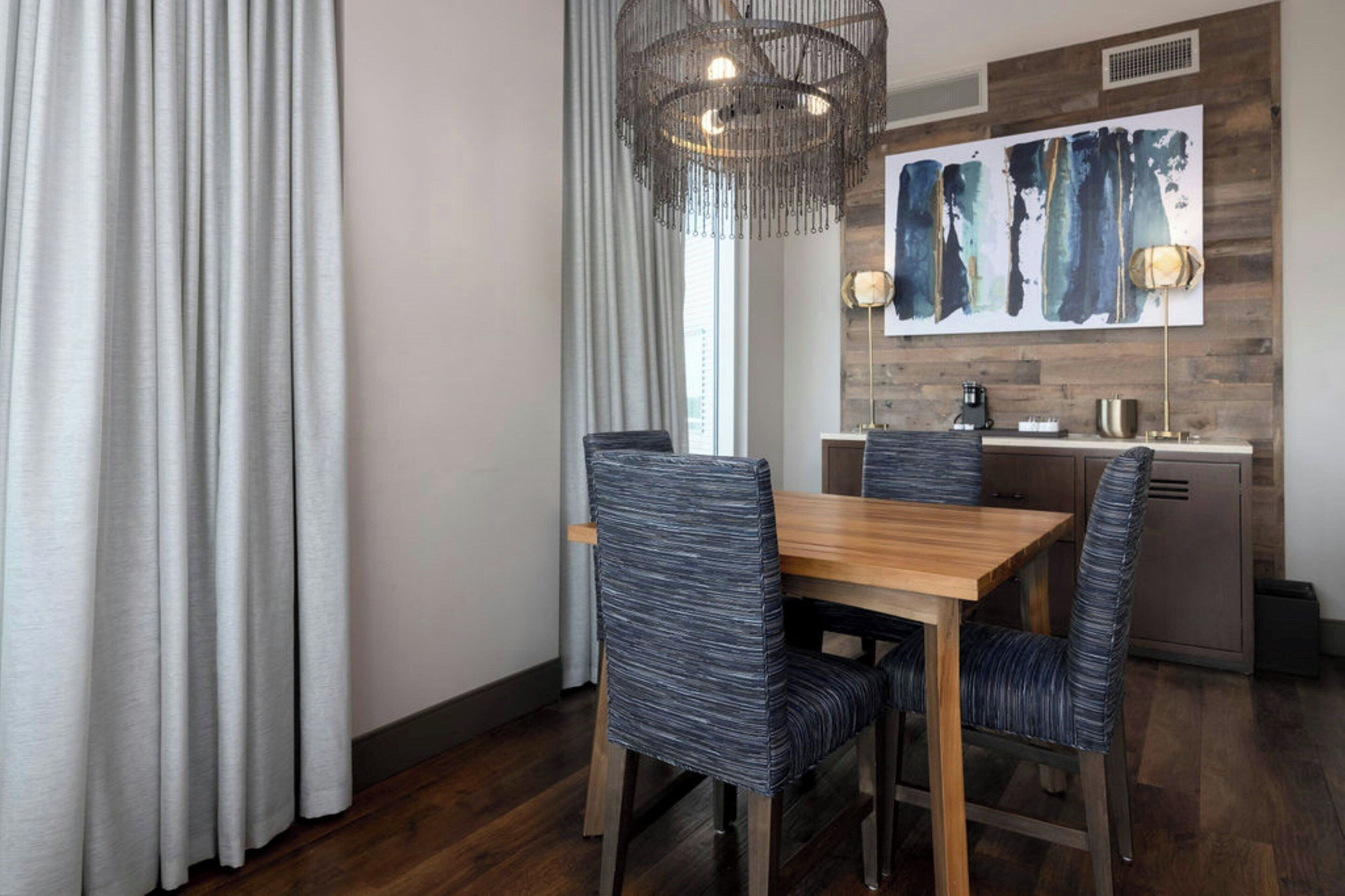 A dining area with a wooden table and chairs, a chandelier, and a kitchen in the background.
