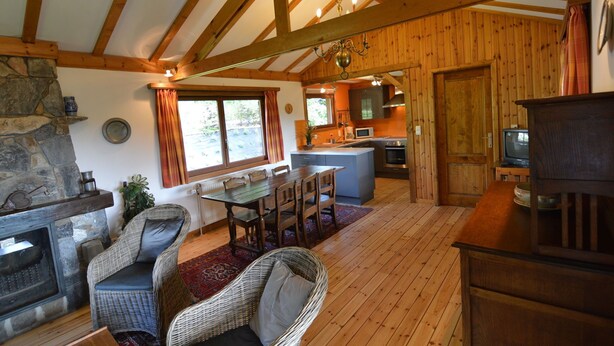 A cozy cabin interior with a stone fireplace, wooden dining table, wicker chairs, a kitchen area, and a television.
