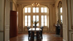 Une salle à manger avec une grande table en bois, des chaises, un lustre et de grandes fenêtres.