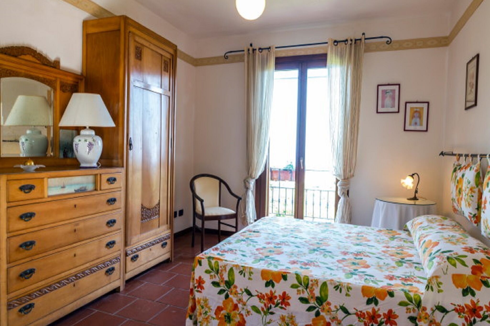 A bedroom with a bed, a wooden dresser, a chair, and a balcony with curtains.