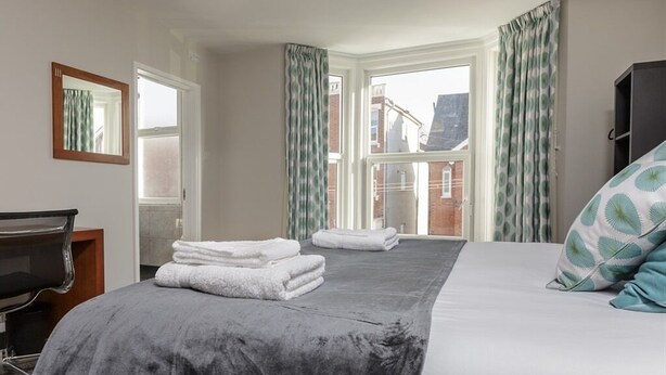 Quarto de hotel com cama, escrivaninha, cadeira e janela com cortinas.