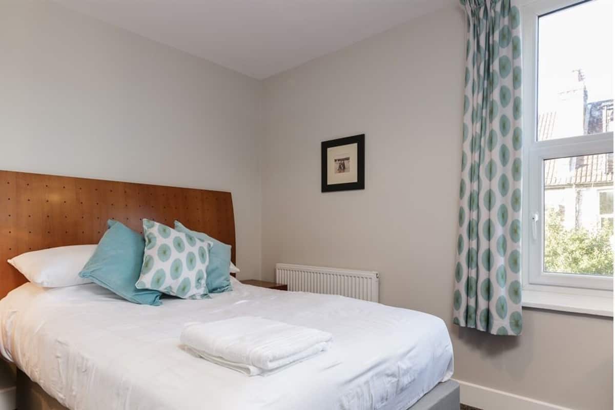 A neatly made bed with white bedding and a few patterned pillows, a wooden headboard, a framed picture on the wall, and a window with curtains.