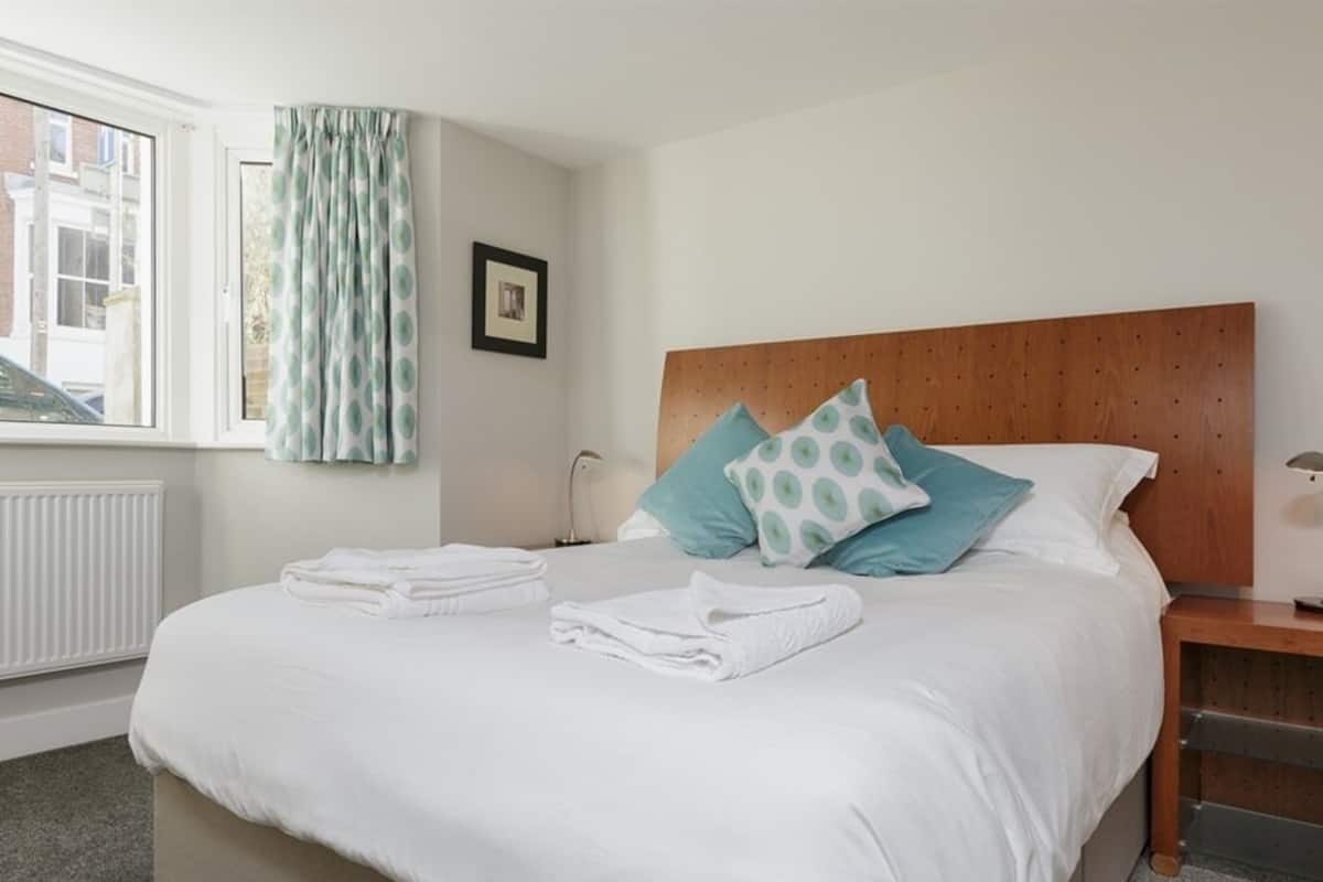 A neatly made bed with white linens and two pillows, a wooden headboard, a bedside table with a lamp, and a window with curtains.