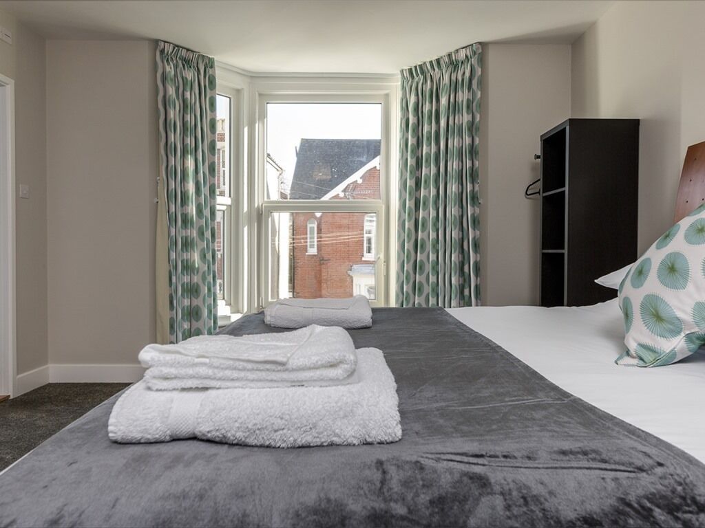 Ett hotellrum med en stor säng, grå sängkläder, en svart garderob och utsikt över en byggnad med tegelstensfasad genom fönstret.