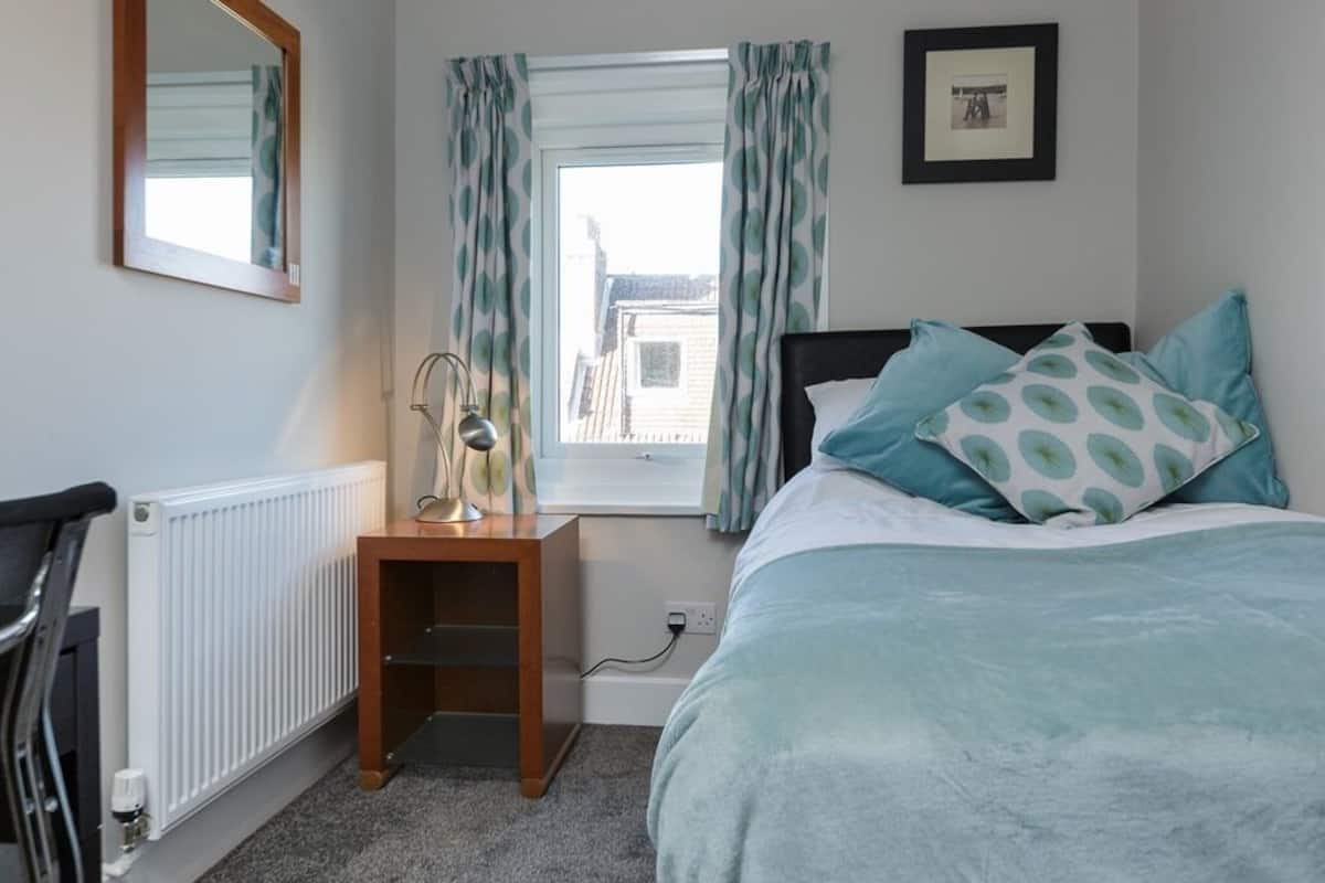 A bedroom with a bed, a nightstand, a chair, a radiator, a window with curtains, and a framed picture on the wall.