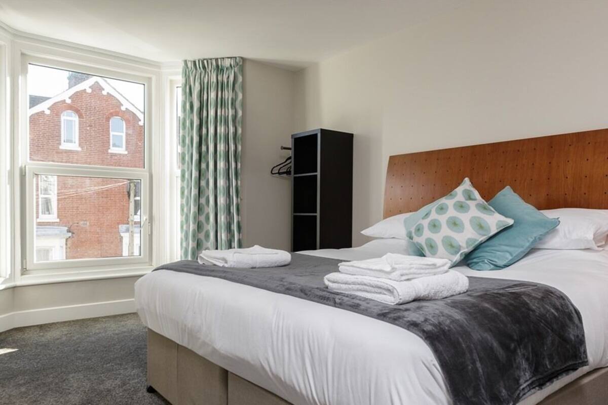 A neatly made bed with a grey and white duvet, a wooden headboard, and two pillows with a leaf pattern. A black wardrobe is visible to the left of the bed.