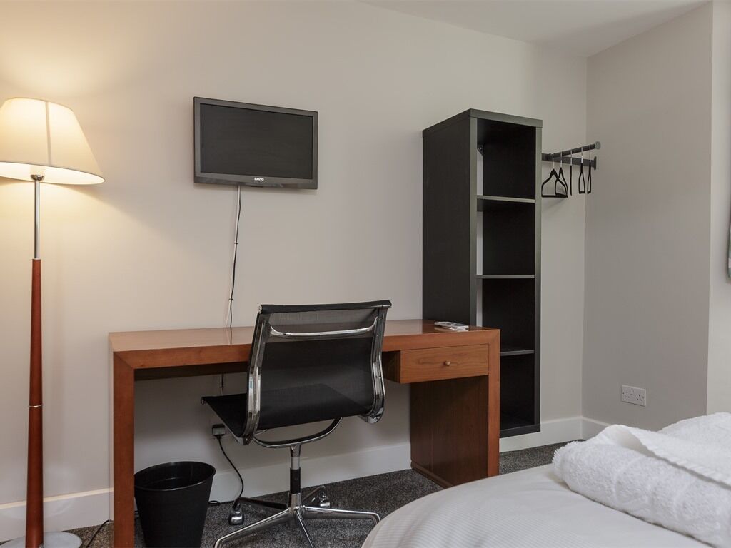 A hotel room with a wooden desk, a black office chair, a television mounted on the wall, a wardrobe, and a bed with white bedding.