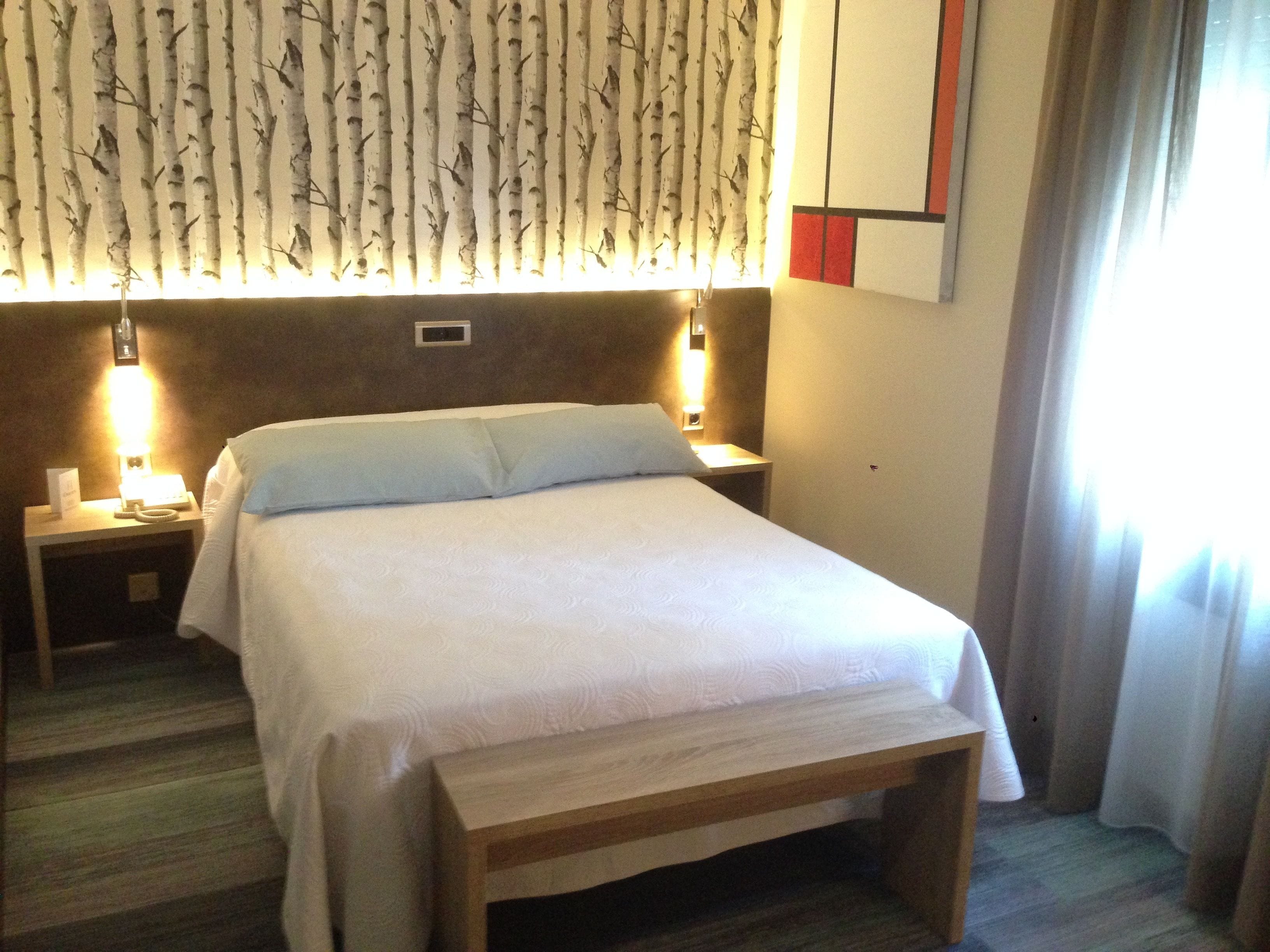 A hotel room with a bed, bedside tables, a nightstand, a wooden bench, a wall with a patterned design, and a window with curtains.