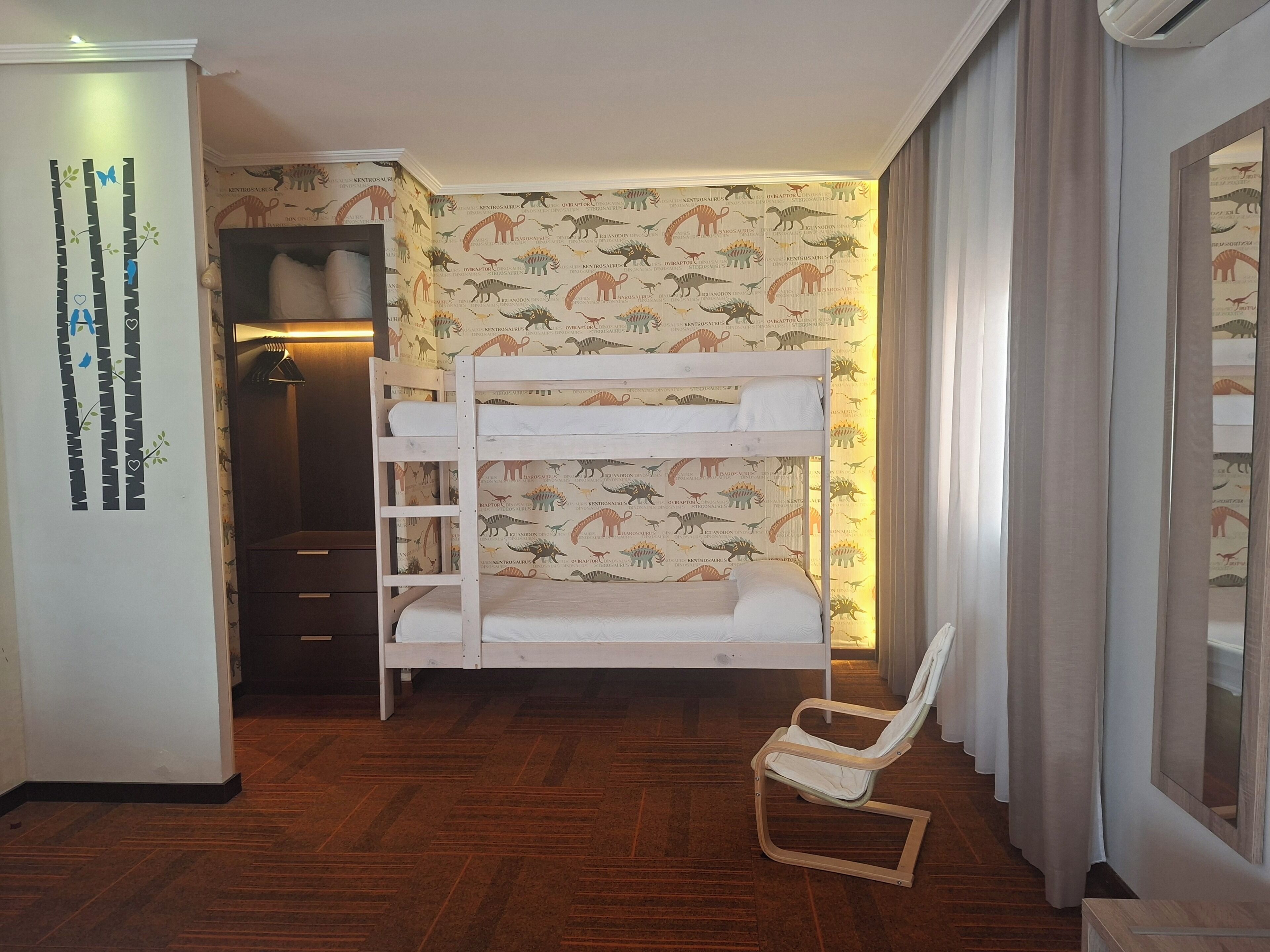 Una cama litera con papel tapiz con motivos de dinosaurios, una silla de madera y un armario con espejo.
