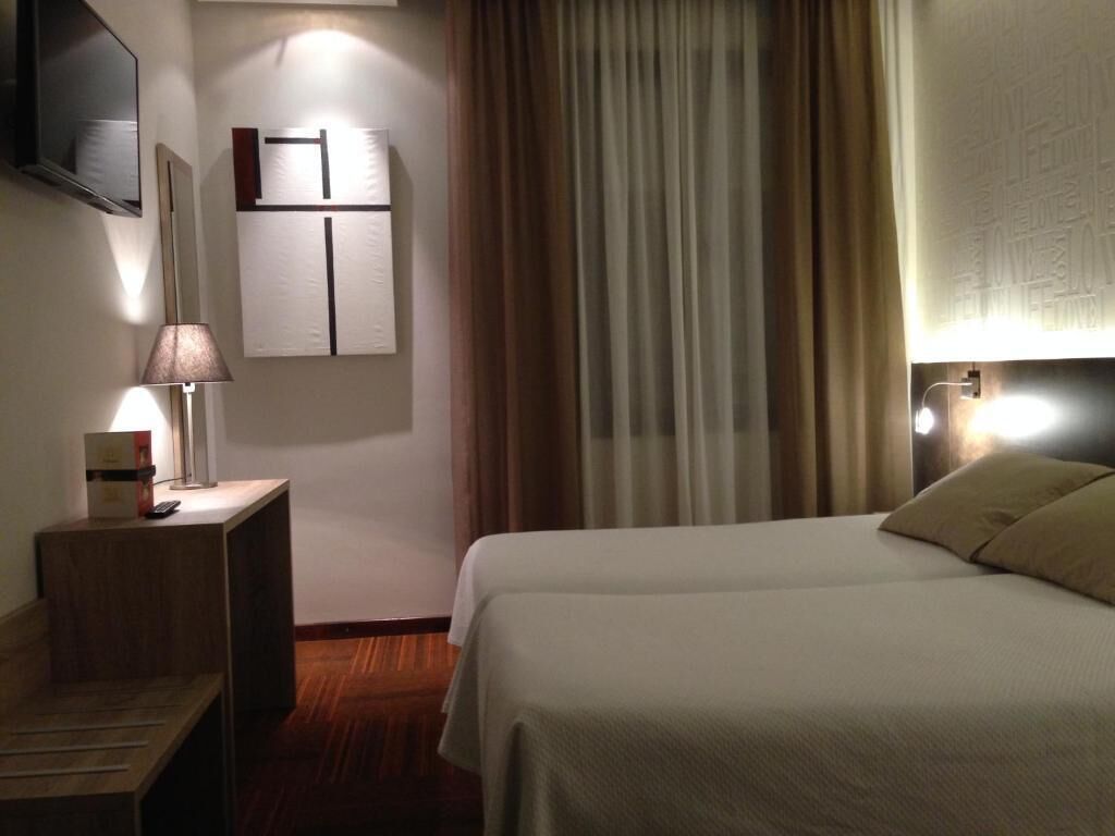 A hotel room with a bed, a nightstand with a lamp, a mirror, and a wall-mounted cabinet.