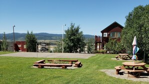 A picnic area with wooden benches and picnic tables, surrounded by a grassy field and a building with a red roof. - Wolf Den Log Cabin Motel and RV Park (Etna)