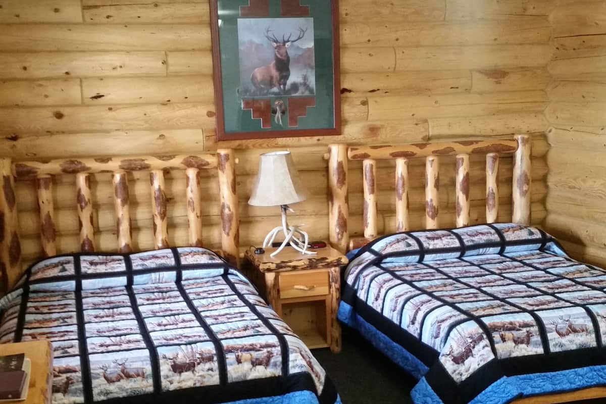 Un dormitorio de cabaña de madera con dos camas, una mesita de noche, una lámpara y un cuadro de un ciervo en la pared.