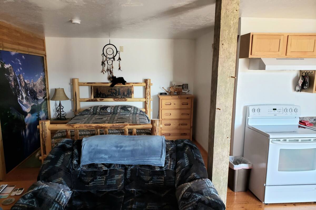 Un dormitorio con una cama, un aparador, una mesita de noche, un atrapasueños, un cuadro y una zona de cocina con estufa.