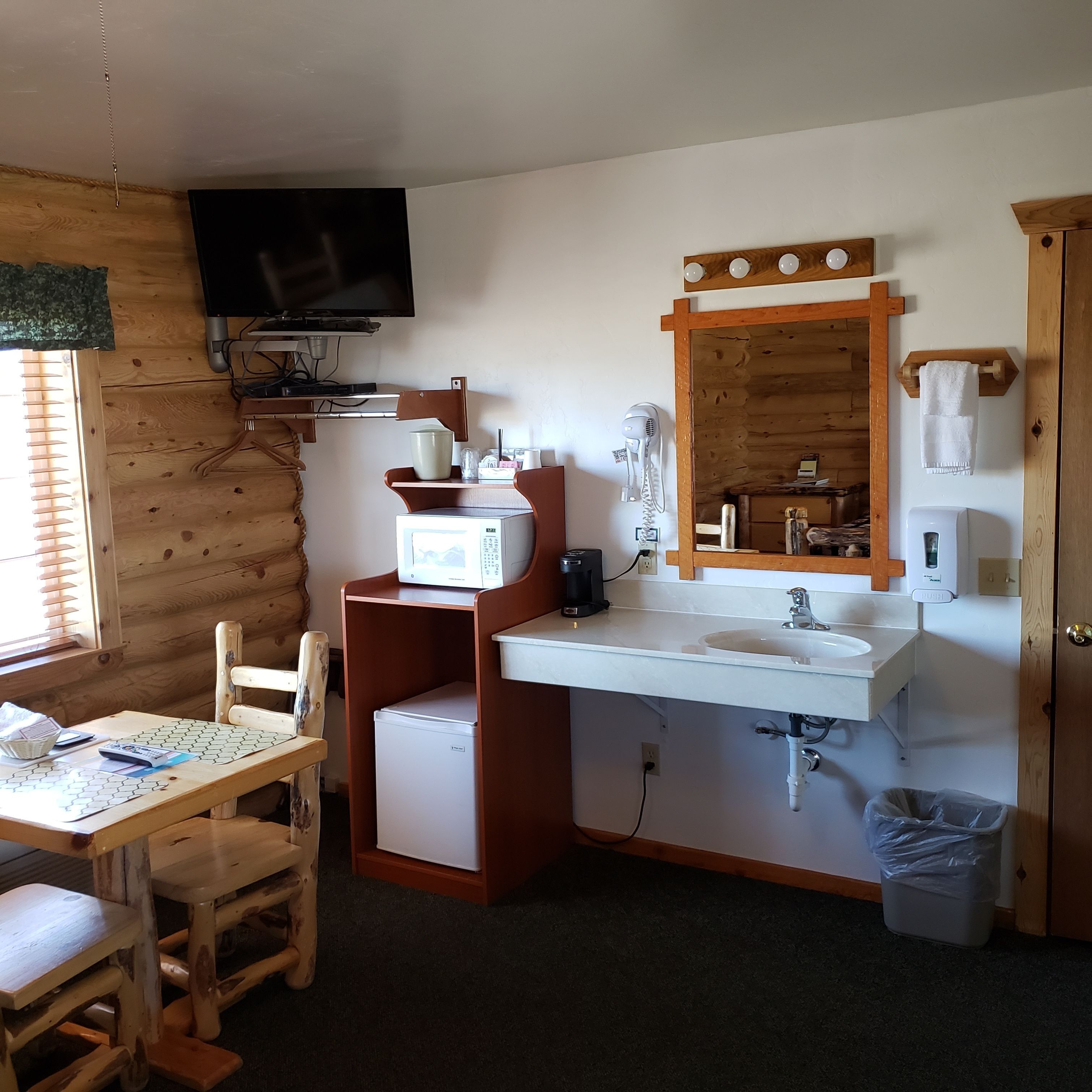 A compact room with a wooden interior, a flat-screen TV mounted on the wall, a microwave, a sink with a mirror above it, a small refrigerator, a trash bin, and a wooden chair.