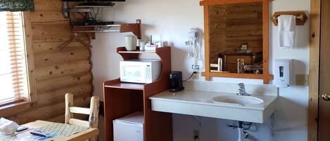 A compact room with a wooden interior, a flat-screen TV mounted on the wall, a microwave, a sink with a mirror above it, a small refrigerator, a trash bin, and a wooden chair.
