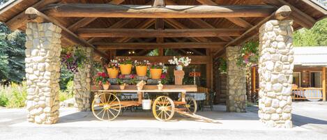 A rustic wooden pavilion with stone pillars, a wooden bench, and a cart with potted plants.