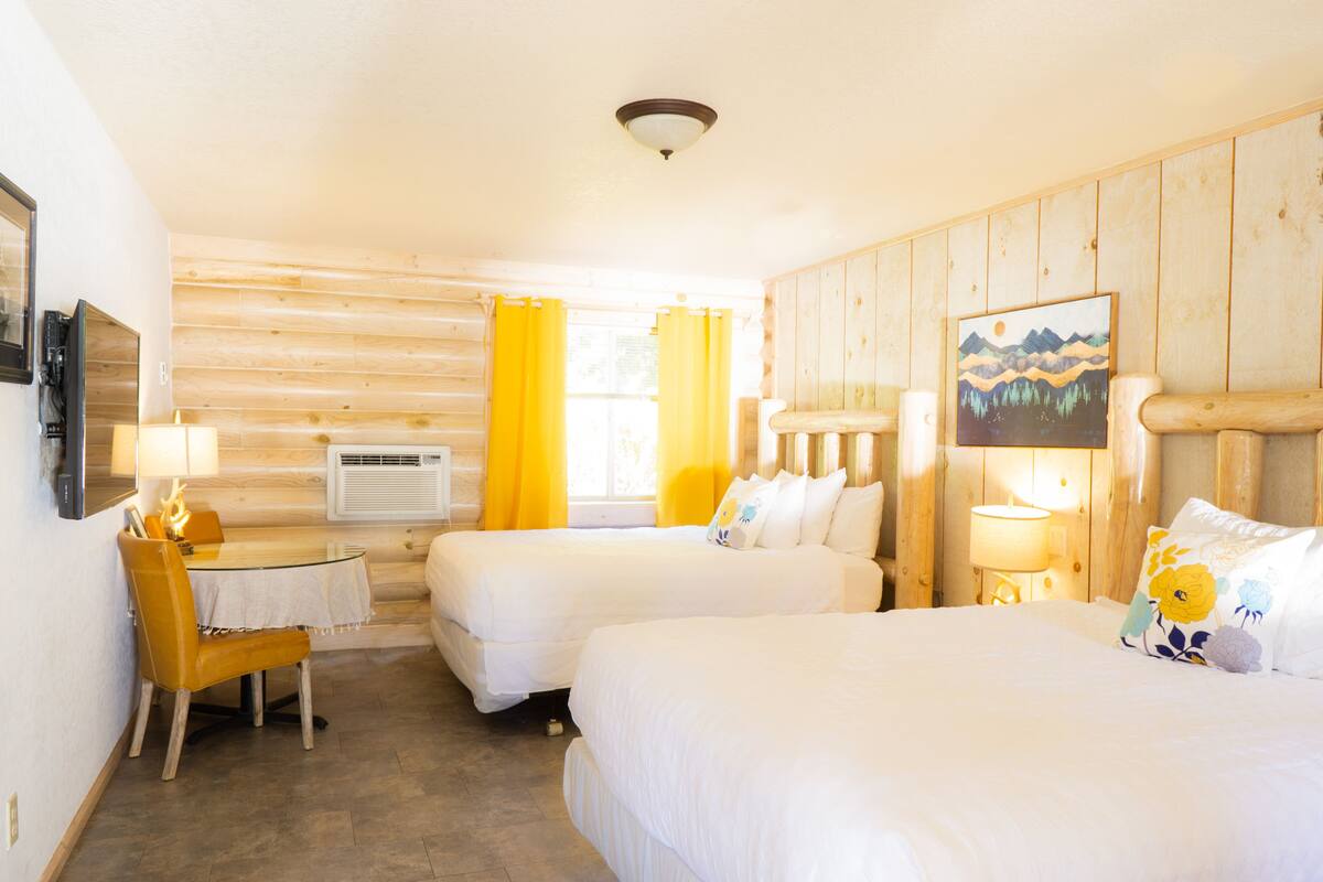 A room with two beds, a desk, and a chair. There is a painting on the wall and a window with curtains.