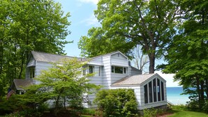 A white house with a large bay window, surrounded by lush greenery and a view of the ocean. - Good Harbor Bay Estate, Lake Michigan (Lake Leelanau)