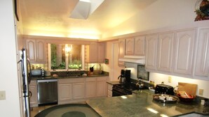 A kitchen with white cabinets, a granite countertop, a stove, a sink, and a window with a view of greenery. - The Weaver House (Glen Arbor)