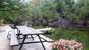 A wooden deck with picnic tables overlooking a calm body of water surrounded by dense greenery. - The Weaver House (Glen Arbor)