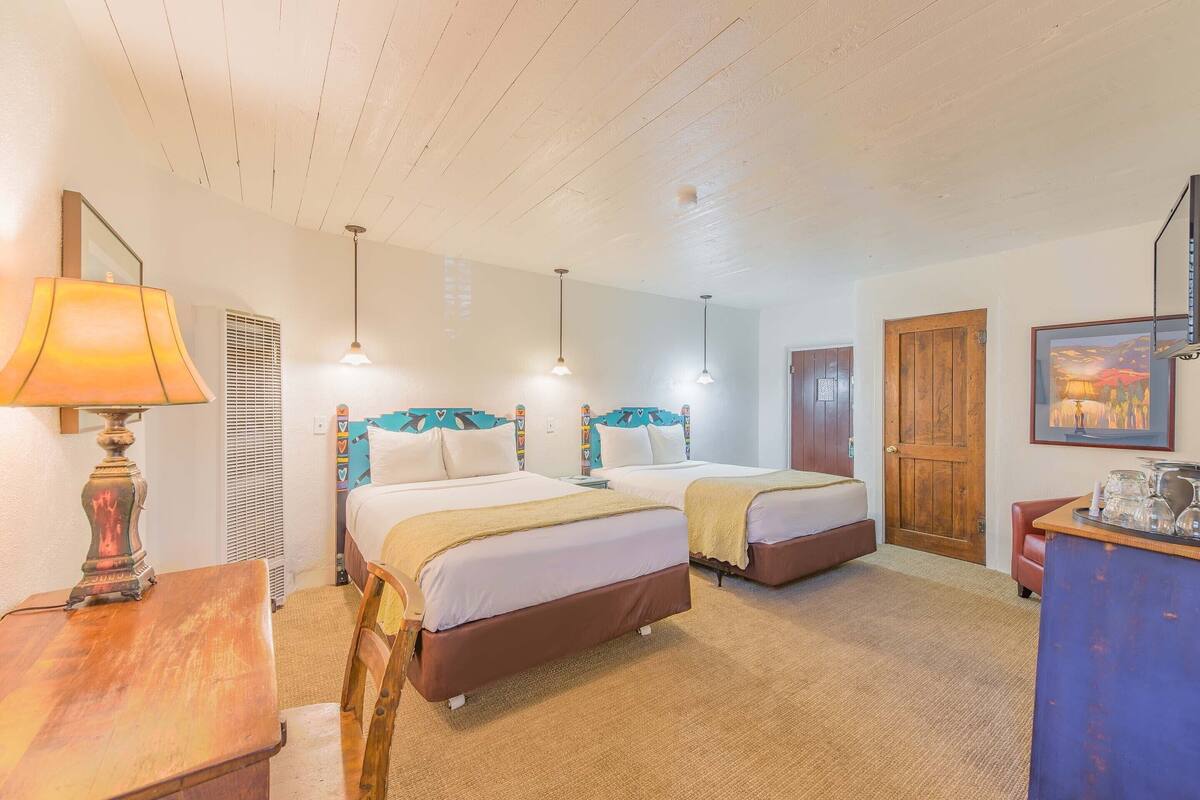 A hotel room with two beds, a wooden desk, a lamp, a door, and a painting on the wall.