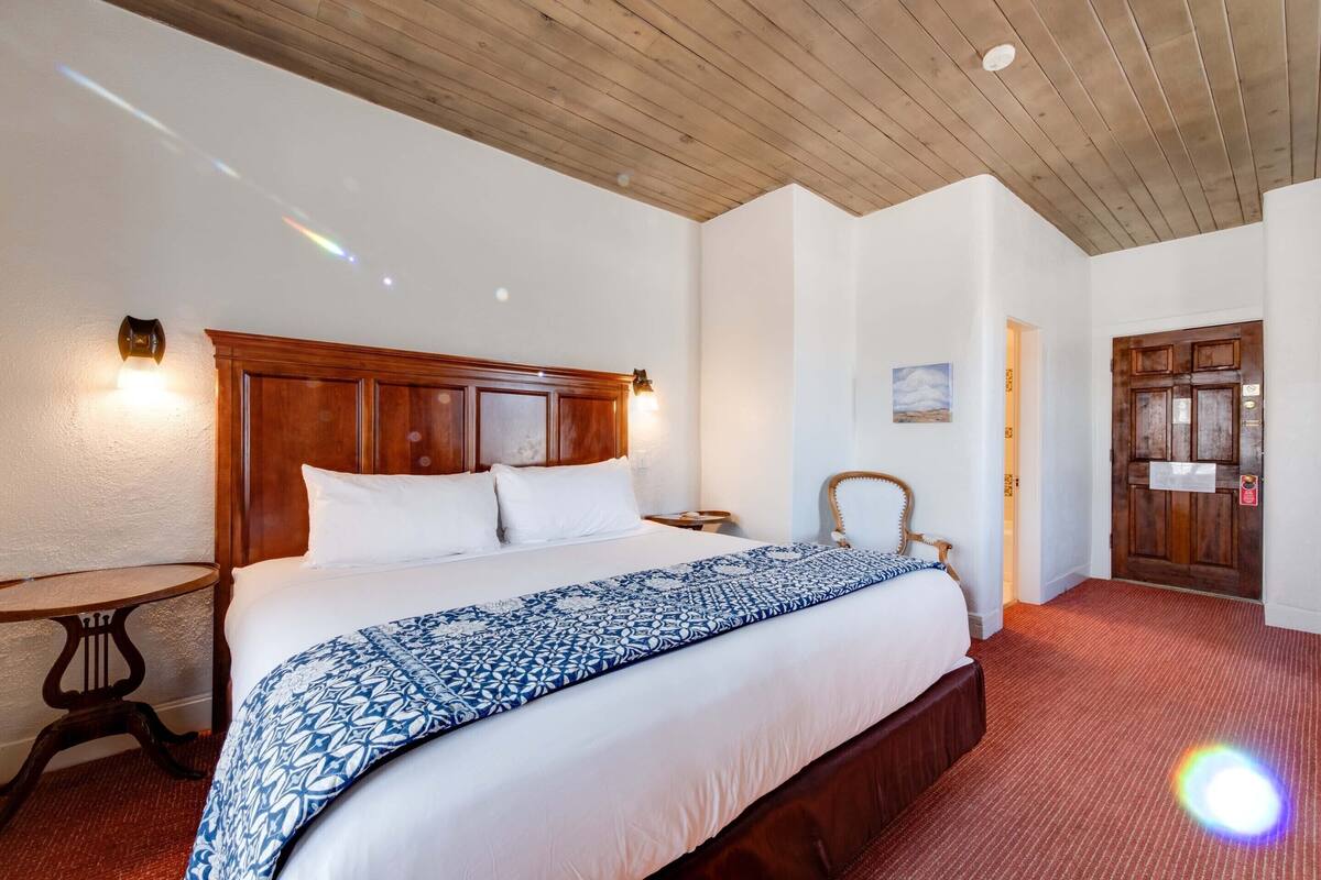 A hotel room with a wooden headboard, a large bed with a patterned bedspread, a small round table, and a door with a sign.